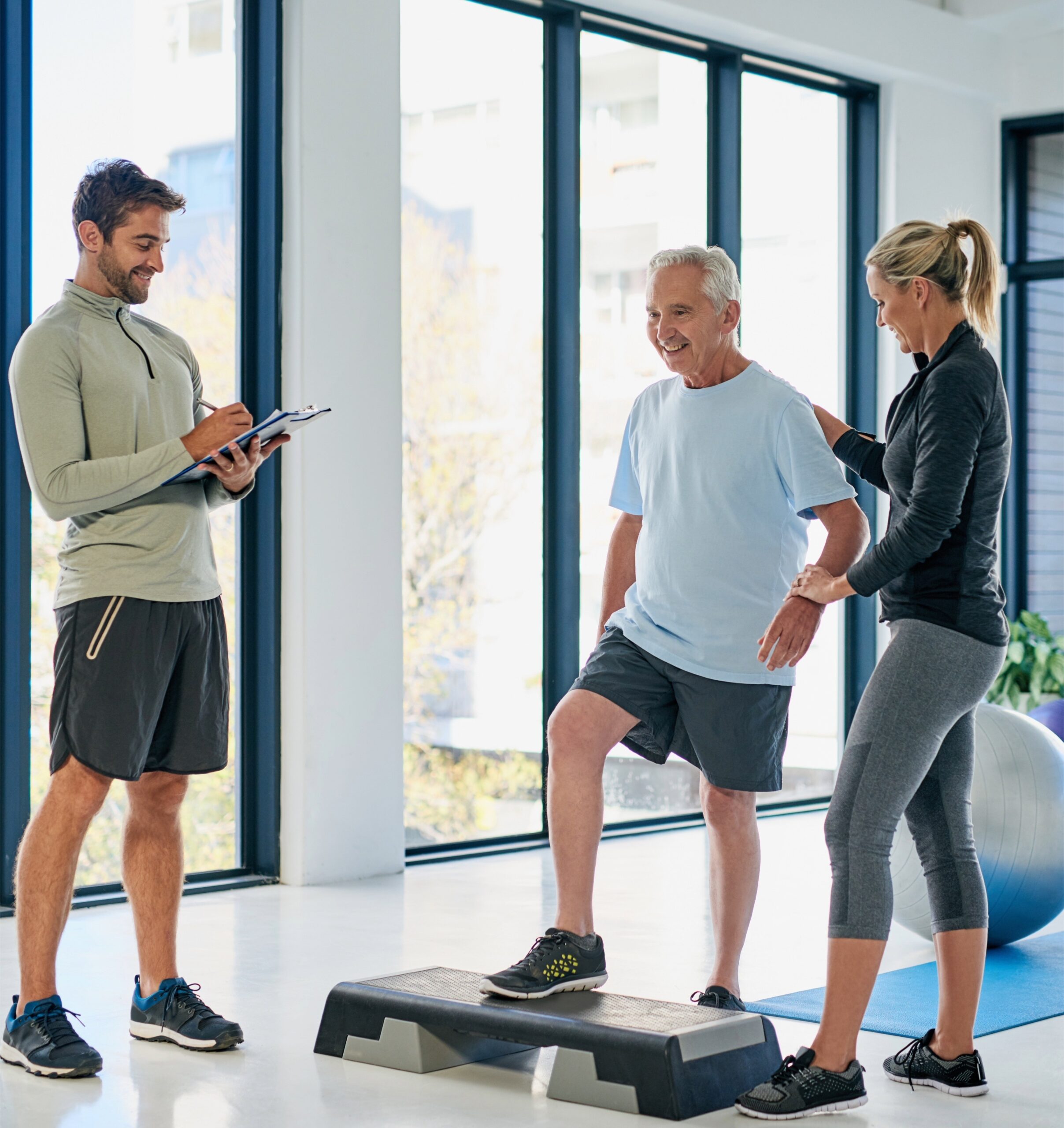 Physiotherapist with a clipboard and a colleague working with a smiling older man in a bright clinic